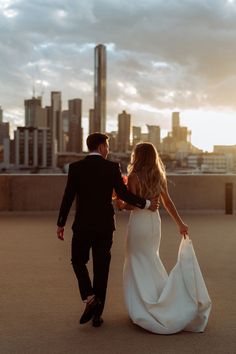 a bride and groom walking together in front of the city skyline at sunset, holding each other's hands