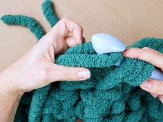 two hands are crocheting the ends of a green piece of cloth with a blue button