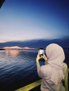 a woman wearing a white hijab is looking at the water with her cell phone