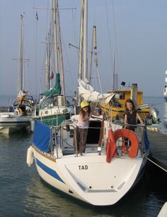 two people standing on the deck of a sailboat docked at a dock with other boats in the background