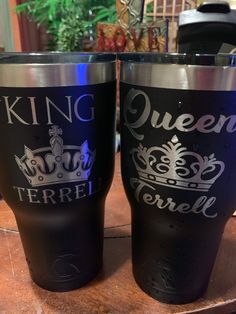 two black tumblers sitting on top of a wooden table next to each other with the words king and queen printed on them