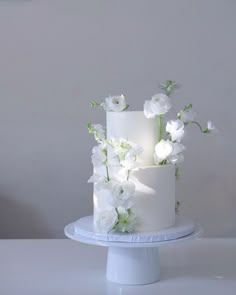 a three tiered cake with white flowers and greenery on the top is sitting on a pedestal