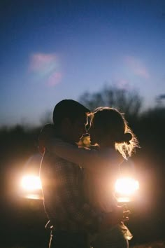 a man and woman embracing in front of their car at night with the headlights on