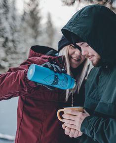 a man and woman standing next to each other in the snow holding coffee mugs