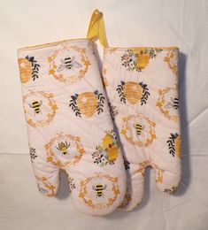 two oven mitts with bees and flowers on them hanging from a yellow ribbon attached to a white background