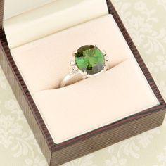 A stunning second hand 18ct white gold green tourmaline & diamond dress ring. An exquisite deep green tourmaline sits at the centre of this gorgeous ring, flanked by a duo of petite diamonds each side. The simple yet stylish setting shows the centre stage gemstone off magnificently whilst the diamonds compliment it perfectly. A part of our second hand gold ring collection. SIZE UK N ½ / US 6 ¾ SPECIFICATION Tourmaline carat weight approx. 5 carats Face dimensions. 11.5 x 14.5 mm Weight. 5.3 gram Elegant Tourmaline Diamond Ring With Accent Stones, Green Tourmaline Jewelry With Halo Setting, Elegant Tourmaline Promise Ring, Elegant Tourmaline Wedding Ring, Elegant Green Topaz Ring In Sterling Silver, Green Tourmaline Wedding Rings, Elegant Green Topaz Ring, Round Cut, Elegant Green Topaz Round Cut Ring, Elegant Green Topaz Ring With Diamond Accents