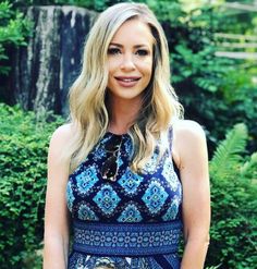 a woman standing in front of some bushes and trees wearing a blue dress with an intricate pattern on it