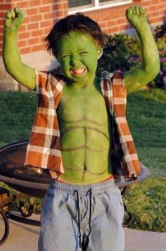 a young boy with green paint on his body and arms, standing in front of a fire pit