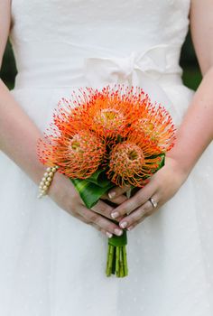 a woman in a white dress holding a bouquet of orange flowers and greenery on her wedding day