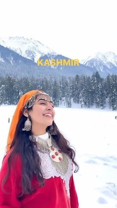 a woman standing in the snow wearing a red dress and headdress with mountains in the background