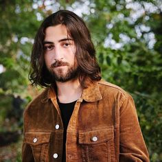 a man with long hair and beard standing in front of trees wearing a brown jacket