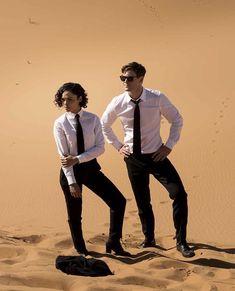 two men in white shirts and black ties standing on sand dunes