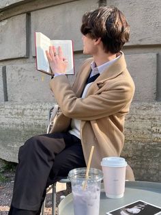 a man sitting on a chair reading a book and holding a drink in his hand