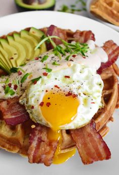 a waffle topped with bacon, an egg and avocado on a white plate