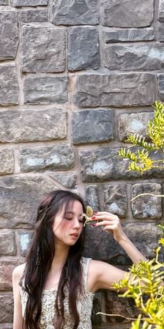 a woman standing next to a brick wall holding a flower in her right hand and looking down