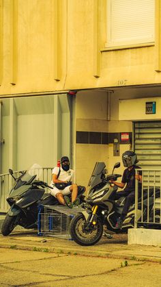 two people sitting on motorcycles in front of a building