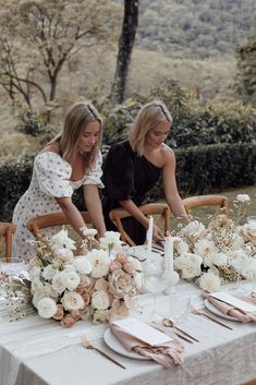 two women sitting at a table with white flowers and place settings in front of them