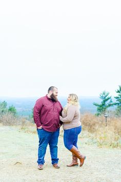 a man and woman standing next to each other on top of a dirt hill with trees in the background