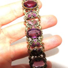 An Edwardian costume bracelet in the Arts and Crafts style made out of cast brass with a spring-ring closure. Each link is made in the design of intertwined curves, decorated with purple enamel leaves, and set with a center faceted purple crystal. This bracelet is in excellent condition and is 18.5 cm long and 2 cm wide. Antique Purple Metal Jewelry, Vintage Adjustable Purple Jewelry, Vintage Purple Bangle Jewelry, Adjustable Vintage Purple Jewelry, Vintage Purple Metal Bracelets, Handmade Vintage Purple Bracelets, Antique Purple Bracelets For Formal Occasions, Vintage Purple Bracelet For Formal Occasions, Purple Bohemian Jewelry For Formal Occasions