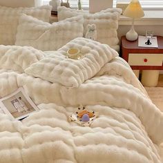 a bed covered in white fluffy blankets next to a night stand with a teddy bear on it