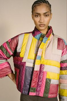 a woman standing in front of a white wall wearing a multicolored quilt jacket