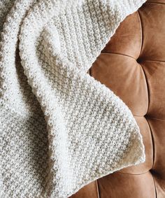 a white knitted blanket sitting on top of a brown leather couch next to a pillow