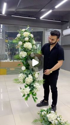 a man standing next to a flower arrangement in a room with white flowers on the floor