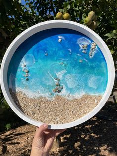 a hand holding up a plate with an image of the ocean in it and sand underneath