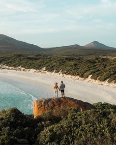 two people standing on top of a rock next to the ocean and hills in the background