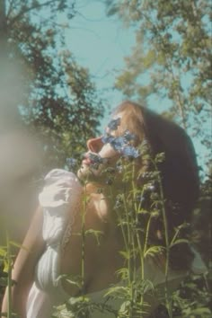 a woman in white dress smelling flowers with trees in the background behind her