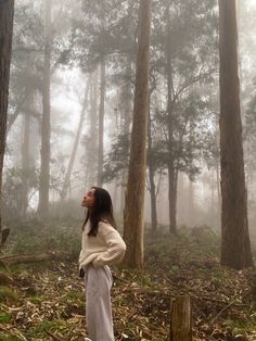 a woman standing in the middle of a forest on a foggy day with her back to the camera