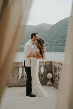 a bride and groom standing on a balcony looking at each other with mountains in the background