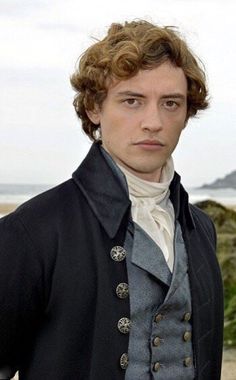 a man with curly hair wearing a black jacket and white shirt standing in front of the ocean