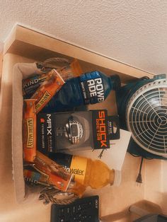 an assortment of sports drinks and snacks in a box on top of a desk next to a fan