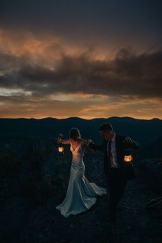 a bride and groom holding lanterns on top of a mountain at sunset with mountains in the background