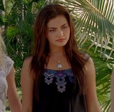 two young women standing next to each other in front of palm trees and plants, one wearing a necklace