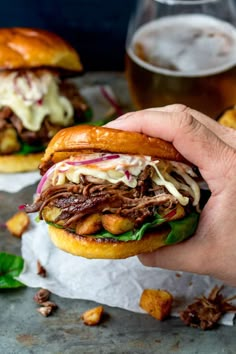 a hand holding a sandwich with meat and cole slaw on it next to a glass of beer