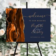 a blue and gold wedding welcome sign on an easel with flowers in the background