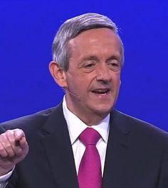 a man in a suit and tie giving a speech on stage with his hand up