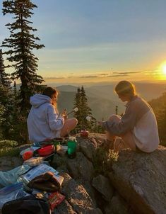 two people sitting on top of a mountain eating food and drinking coffee at sunset with the sun setting behind them