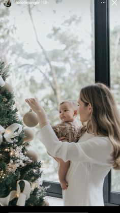 a woman holding a baby in her arms near a christmas tree with ornaments on it