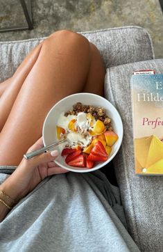 a woman sitting on a couch with a bowl of fruit and a book in front of her