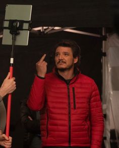 a man in a red jacket is holding a camera and pointing to the side with his finger