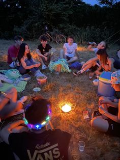 a group of people sitting in the grass around a fire with some lights on it