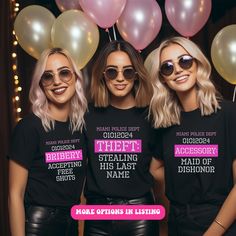 three women wearing black shirts with pink and white letters on them, standing in front of balloons