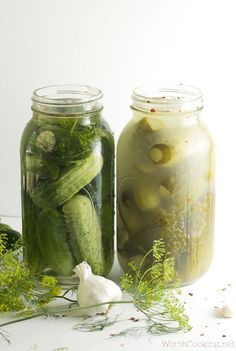 three jars filled with pickles sitting on top of a white table next to garlic and herbs