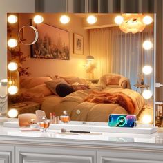 a mirror with lights on it in front of a bed and desk area next to a window