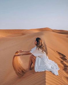 a woman sitting on top of a sand dune in the desert wearing a white dress