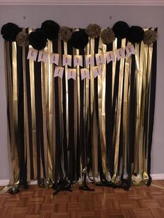 a black and gold birthday party backdrop with paper flowers on the top, hanging by ribbon