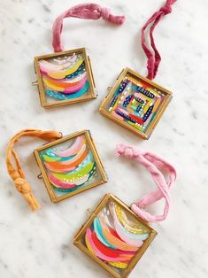 three small square pendants on a marble surface with pink cords and beads hanging from them